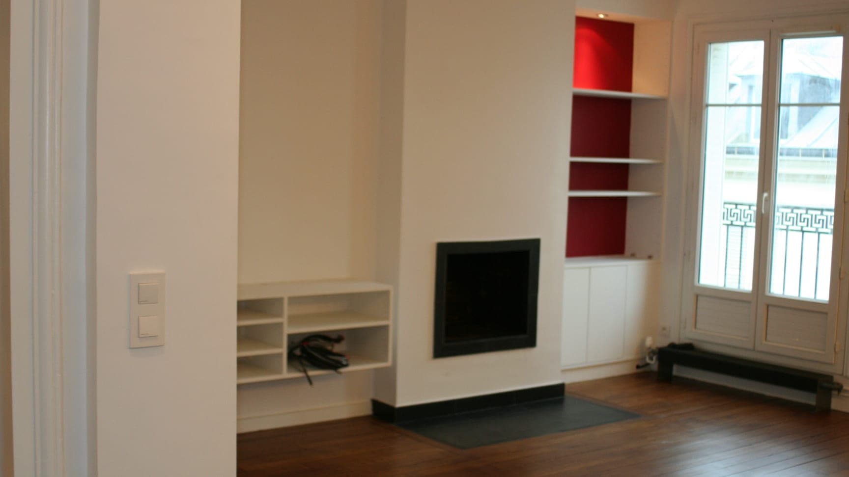 Séjour d'un appartement rénové : cheminée au foyer noir encastré, niches rouges et parquet foncé