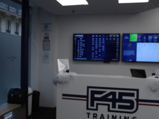 Accueil d'une salle de sport : comptoir blanc au logo F45 Training, deux écrans muraux et plafond lumineux