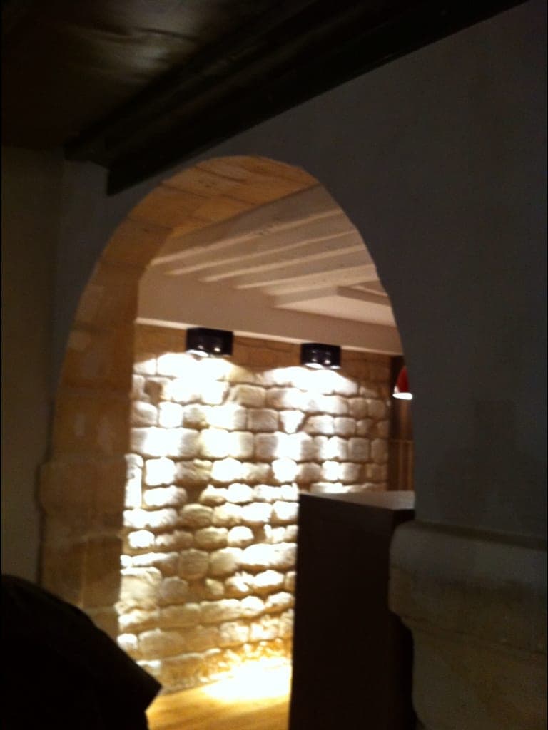 Arche en pierre de taille ouvrant sur un mur de moellons éclairé par des spots, poutres au plafond