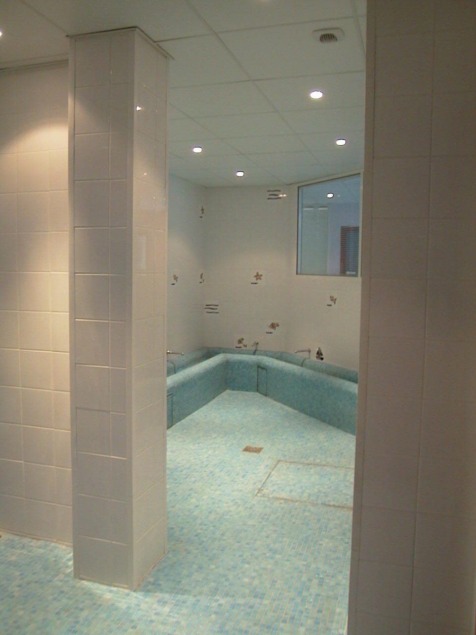 Bassin turquoise vu entre un poteau carrelé blanc et une cloison, faux plafond à dalles et spots
