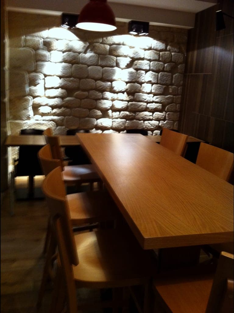 Mur de moellons de pierre en lumière rasante, longue table en bois clair, chaises en bois et suspension rouge