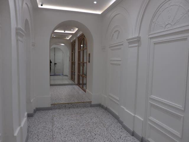 Couloir en enfilade d'arches : panneaux moulurés blancs, granito moucheté, gorge lumineuse, porte vitrée au fond