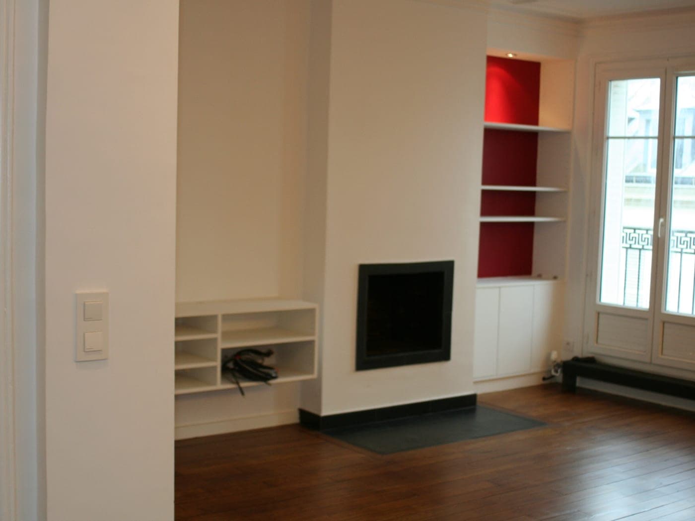 Séjour à parquet foncé : cheminée encastrée à foyer noir, niches à fond rouge et porte-fenêtre sur balcon
