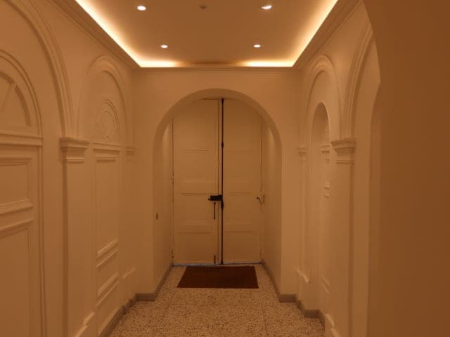 Hall en lumière chaude : arcades et panneaux moulurés, double porte en arc au fond, granito et gorge lumineuse