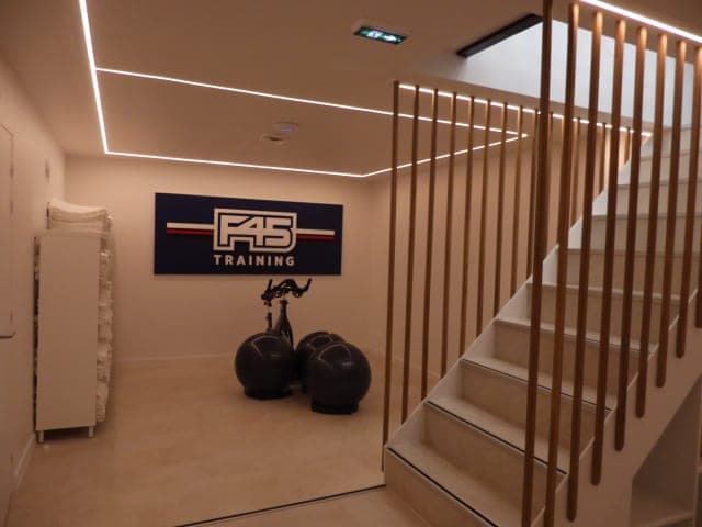 Escalier blanc à garde-corps en lames de bois, lignes lumineuses au plafond, panneau F45 Training au mur