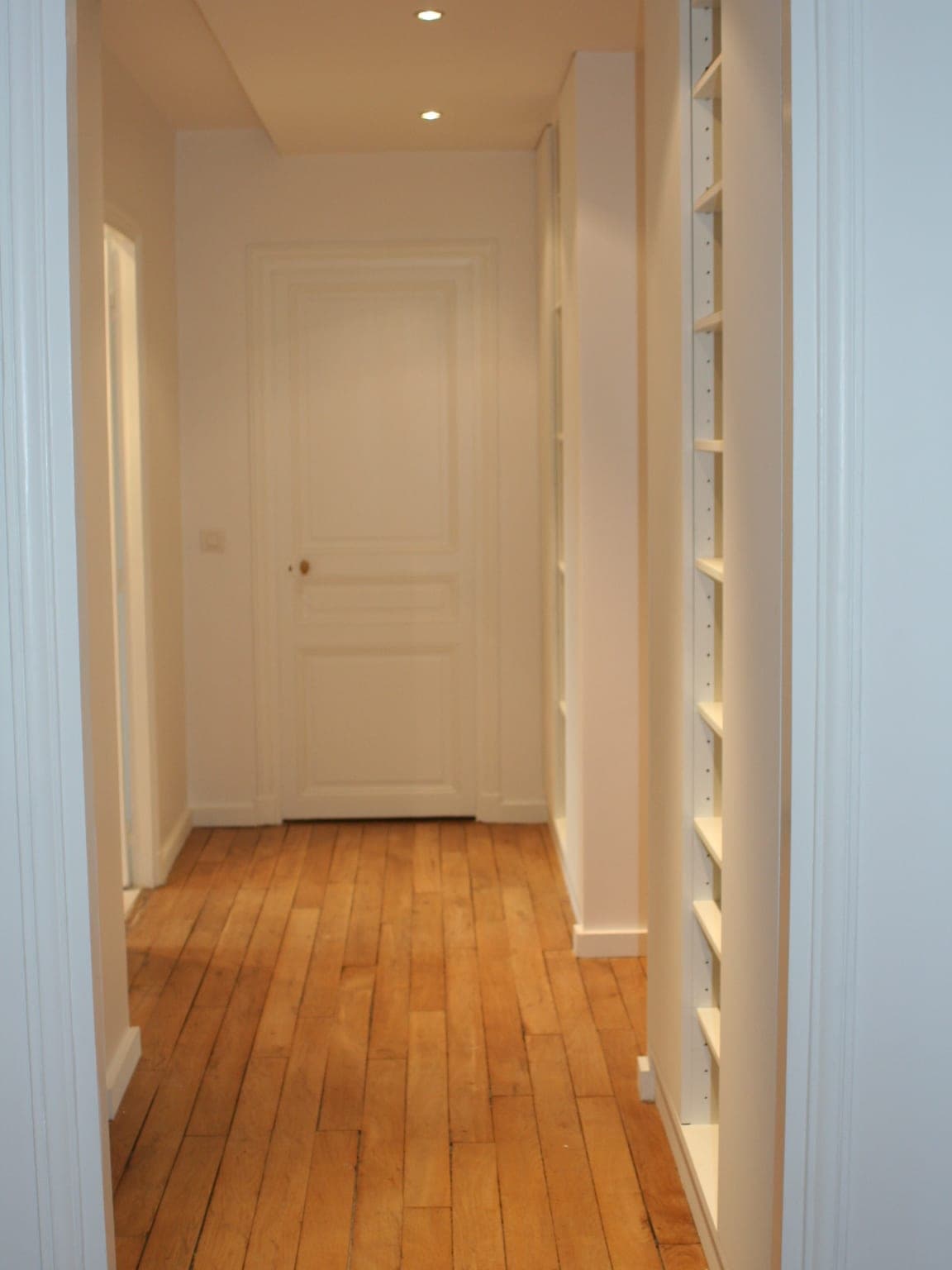Couloir à parquet de chêne en lames, rangements encastrés éclairés et porte moulurée blanche au fond