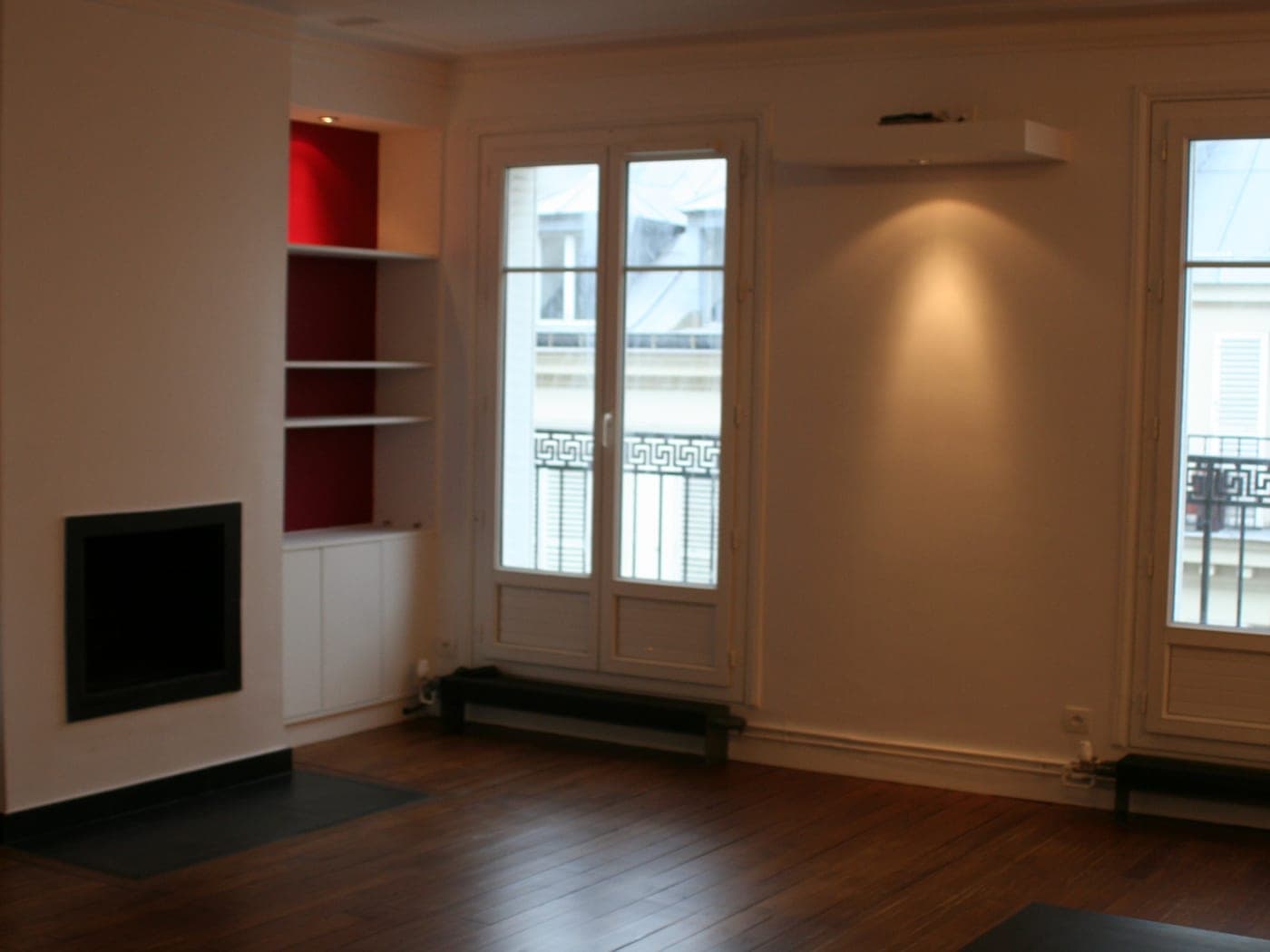 Séjour à parquet foncé : deux portes-fenêtres à balcon, cheminée encastrée et niches à fond rouge, vue large