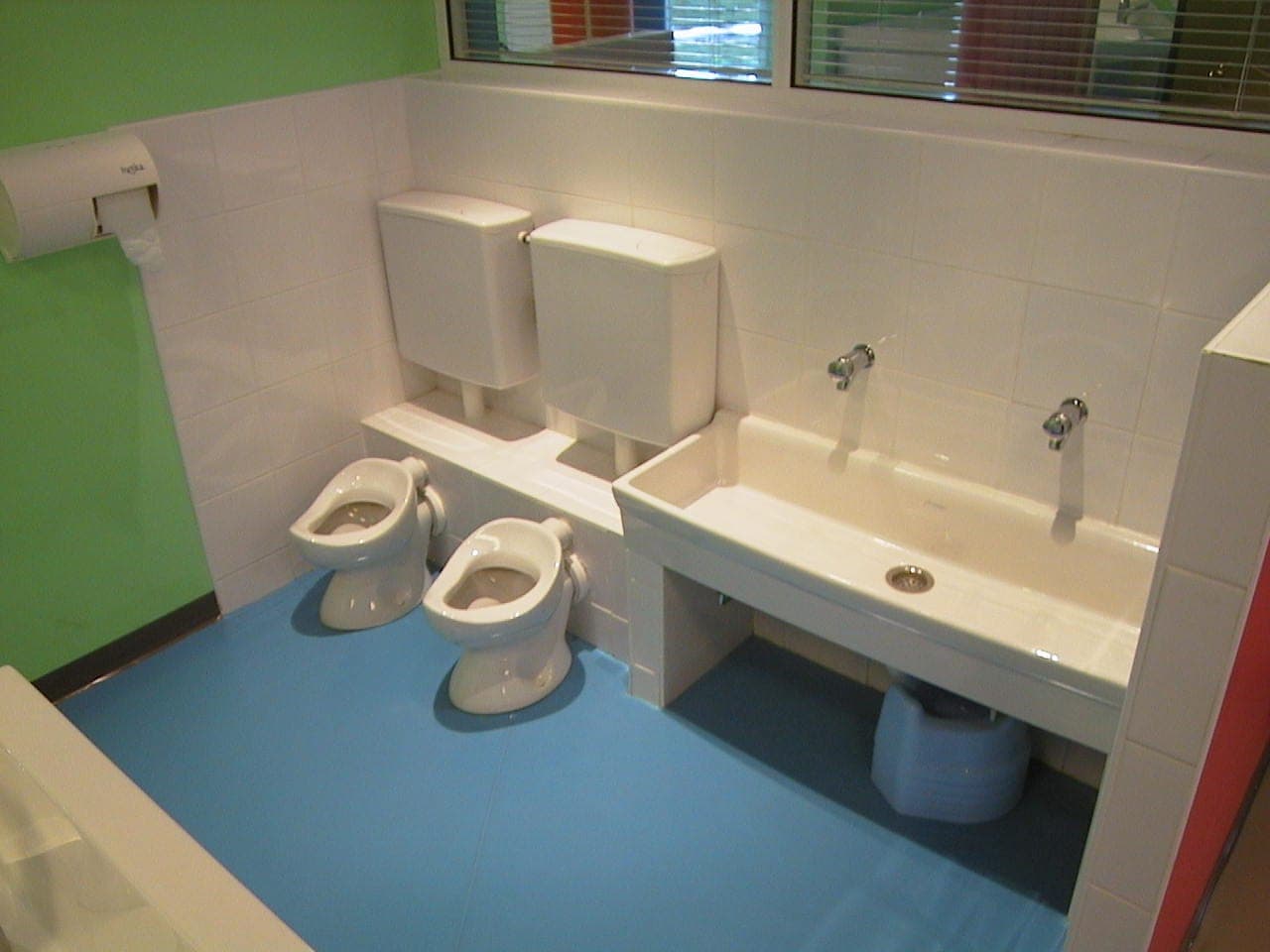 Sanitaires à hauteur d'enfant : petites cuvettes, auge-lavabo carrelée blanche, sol bleu et mur vert