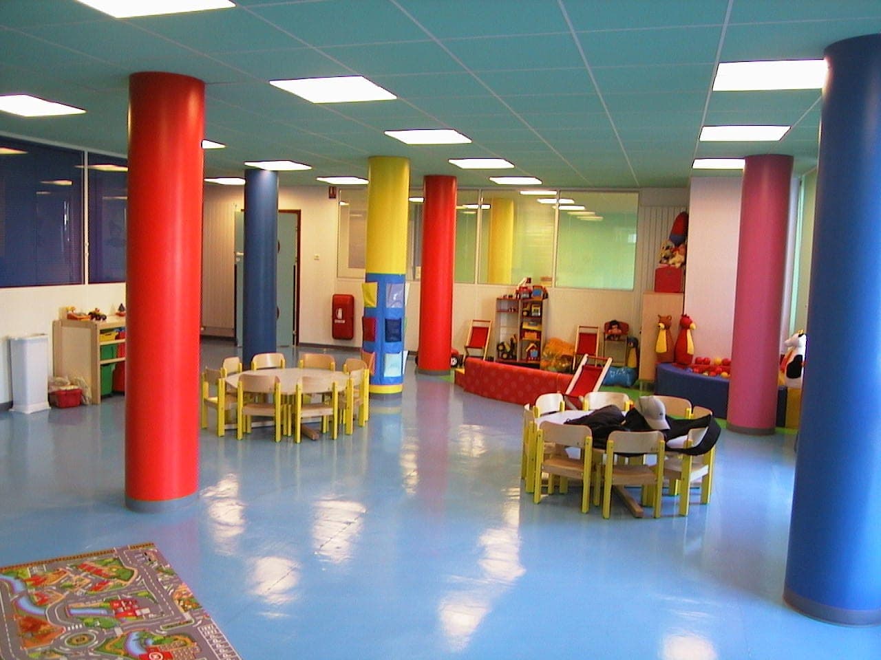 Grande salle aux poteaux ronds peints de couleurs vives, sol souple bleu et petites tables d'enfant