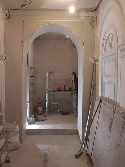 Hall d'immeuble en travaux : arche en plaques de plâtre brutes, gaines pendantes, établi et sac de gravats