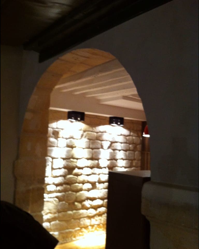 Arche en pierre de taille, mur de moellons éclairé par des spots et poutres au plafond