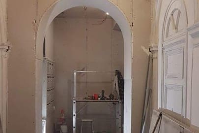 Hall d'immeuble en chantier : arche en plaques de plâtre brutes, gaines électriques pendantes et escabeau