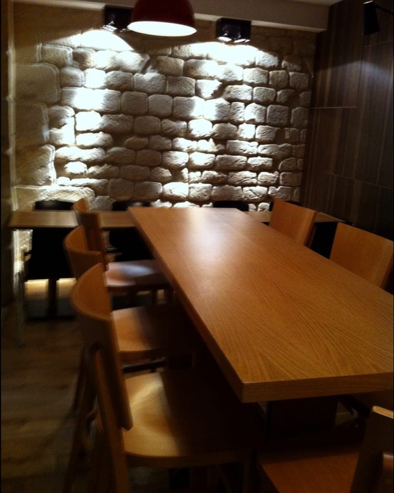 Mur de moellons éclairé en lumière rasante par des spots, longue table en bois clair et chaises