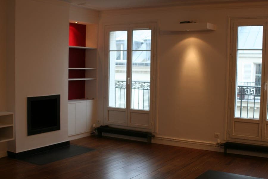 Séjour à parquet foncé : deux portes-fenêtres à balcon, cheminée encastrée et niches à fond rouge