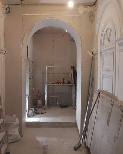 Arche en plaques de plâtre brutes dans un hall en travaux, gaines pendantes et établi au fond